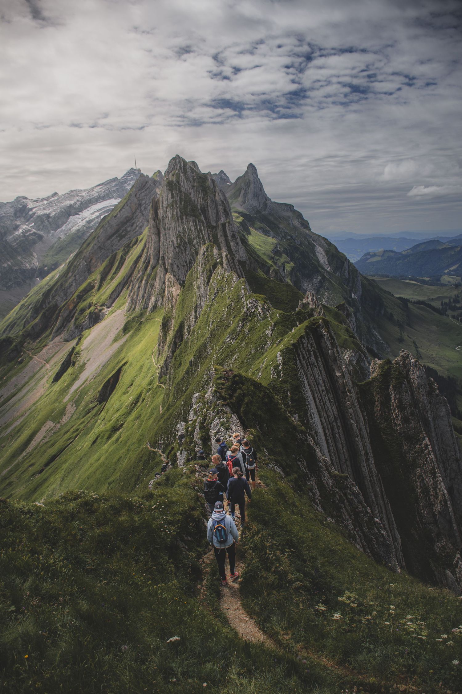 A group of hikers with backpacks trek along a narrow ridge trail toward jagged green mountain peaks.