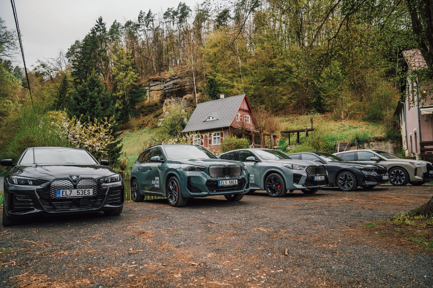Four BMW electric vehicles lined up on a gravel lot with a rustic red cottage and forest in the background.