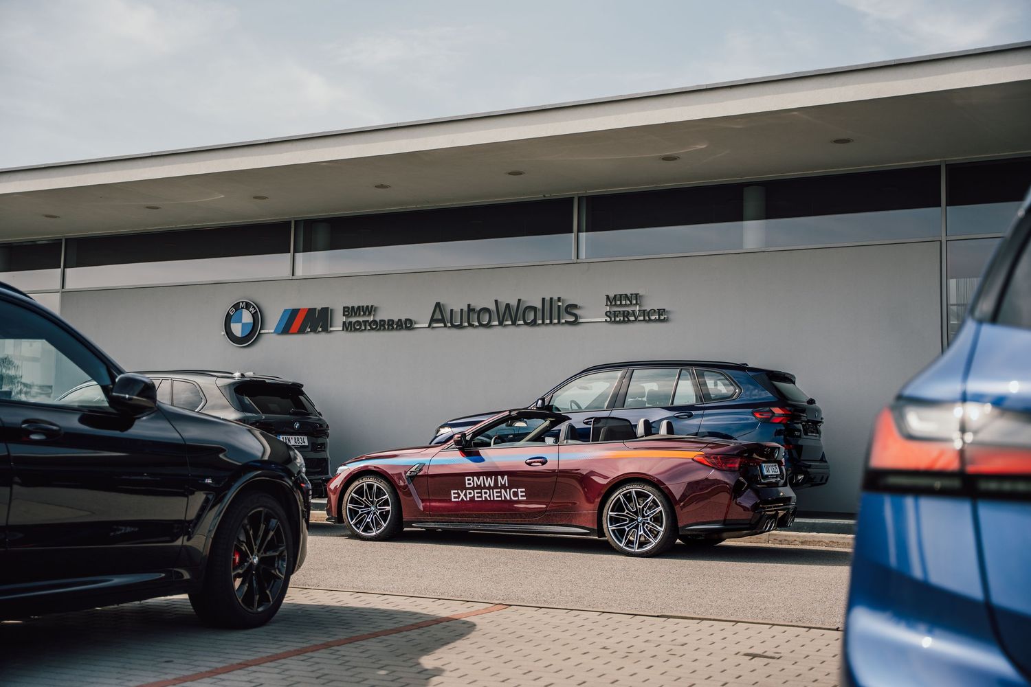 BMW M Experience cars parked outside AutoWallis BMW M dealership on a sunny day.