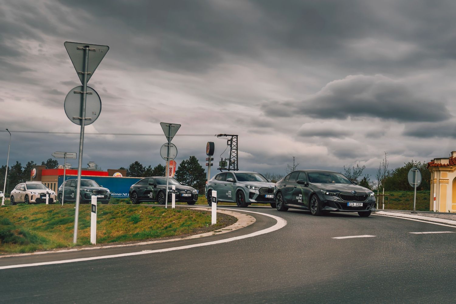 Multiple BMW vehicles gathered at a roundabout intersection under dramatic stormy skies.