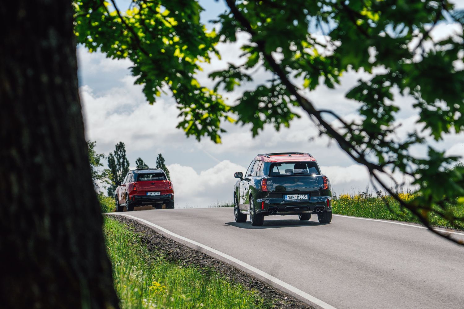 Two MINI Cooper cars driving along a scenic rural road flanked by green fields and trees.