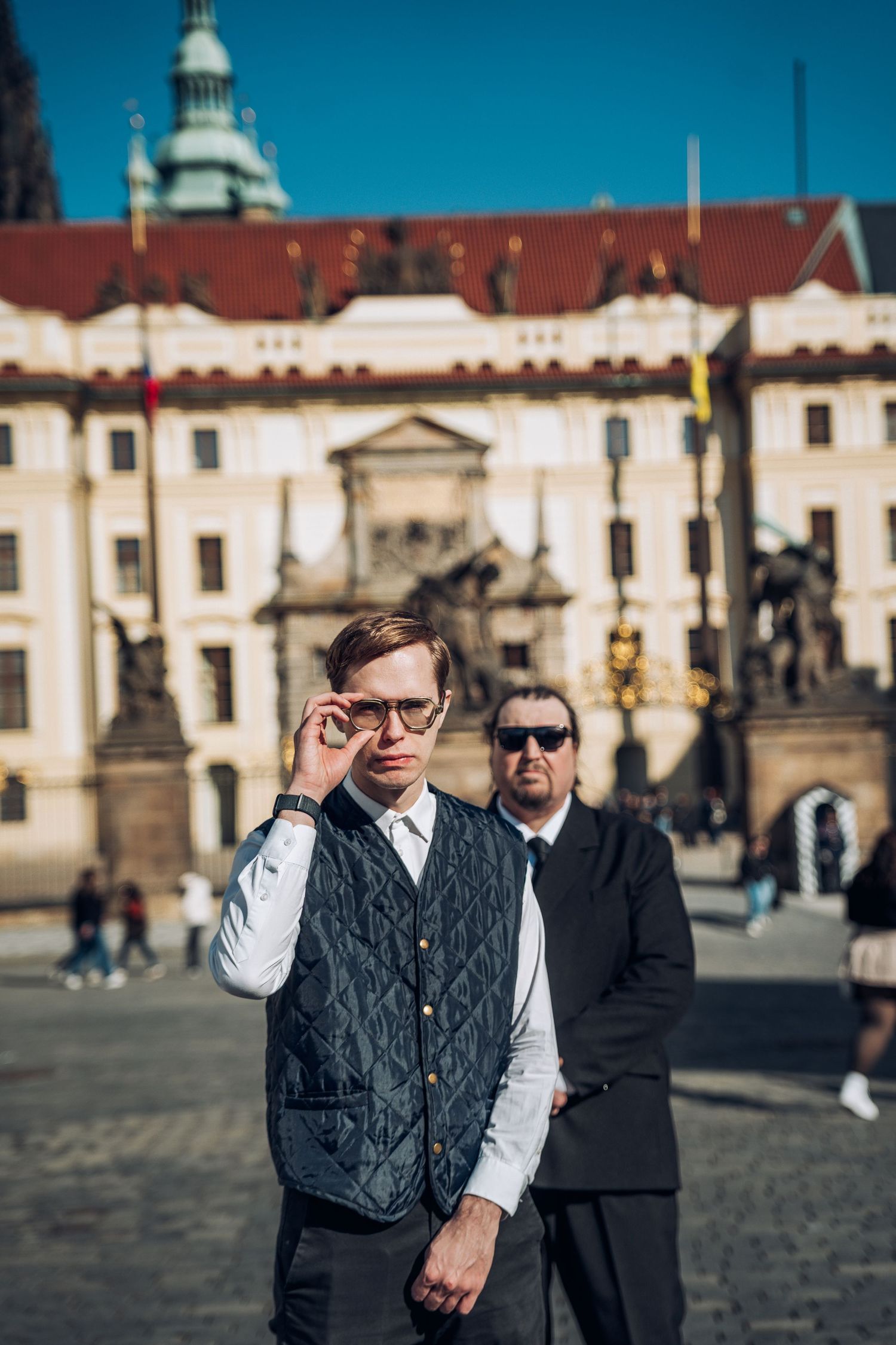 Two men in formal attire pose outdoors near Prague Castle on a sunny day.