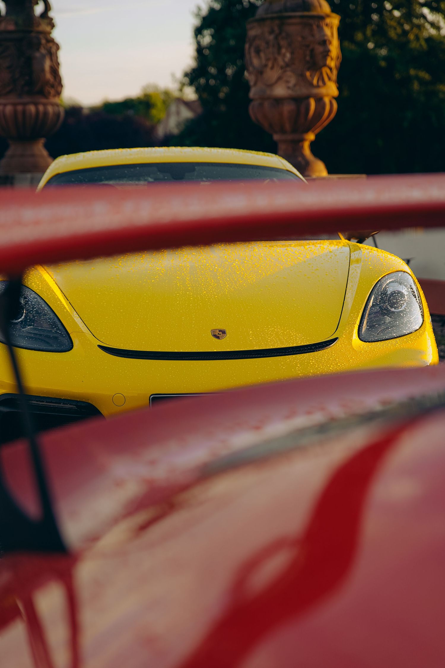 Yellow Porsche 718 Cayman GT4 front end viewed through red car window at golden hour.