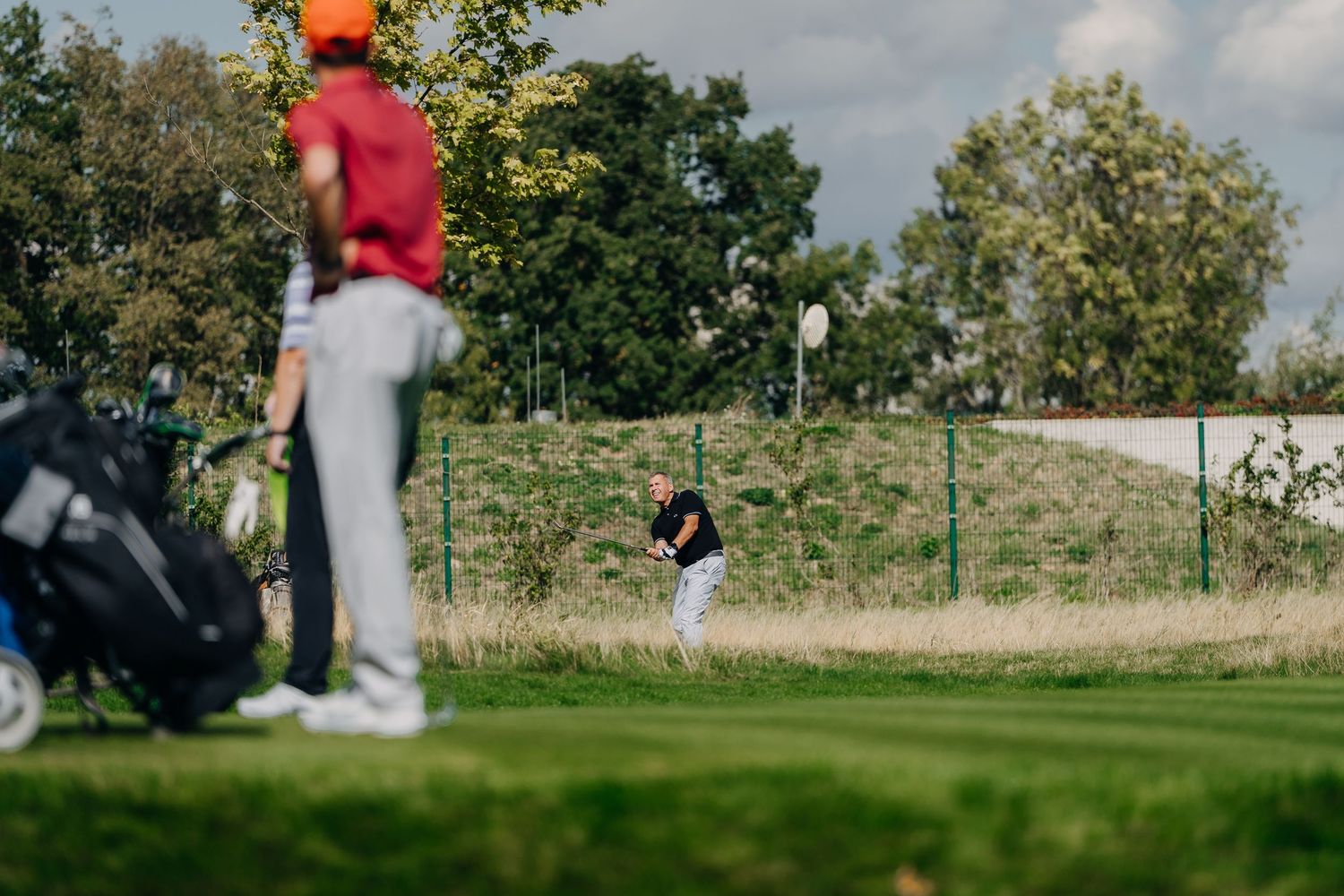Golfer hitting a bunker shot on a golf course while another player watches nearby with a golf bag.
