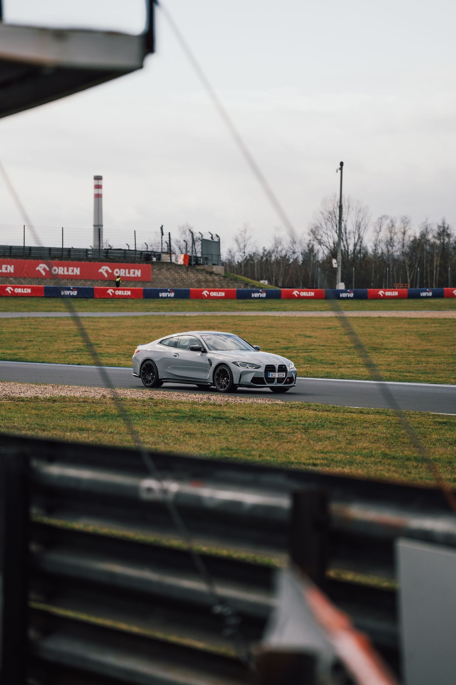 Silver BMW M4 driving on a race track circuit under overcast skies with ORLEN advertising boards visible.