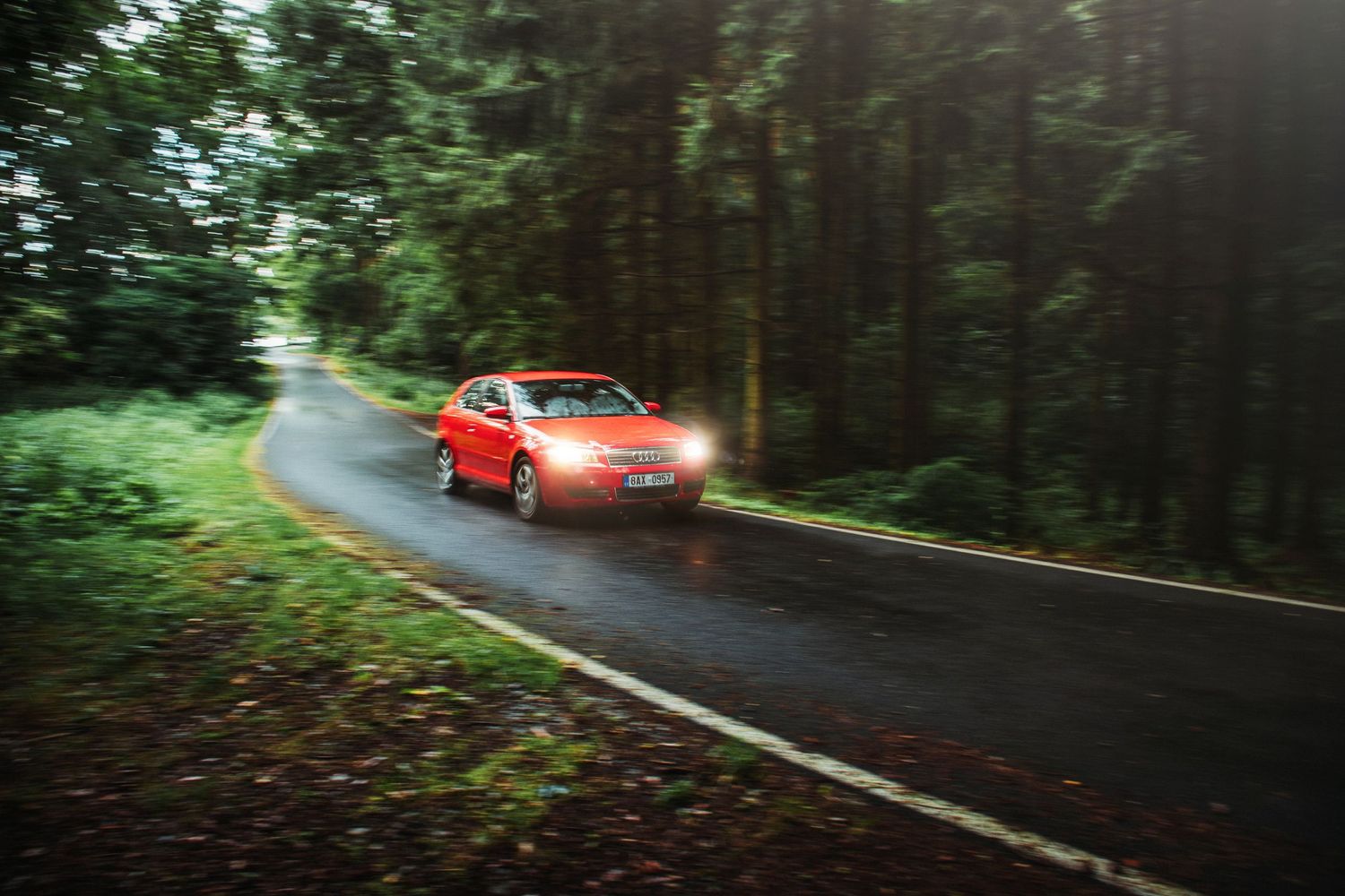 Red sedan speeding through a lush green forest road with motion blur effect.