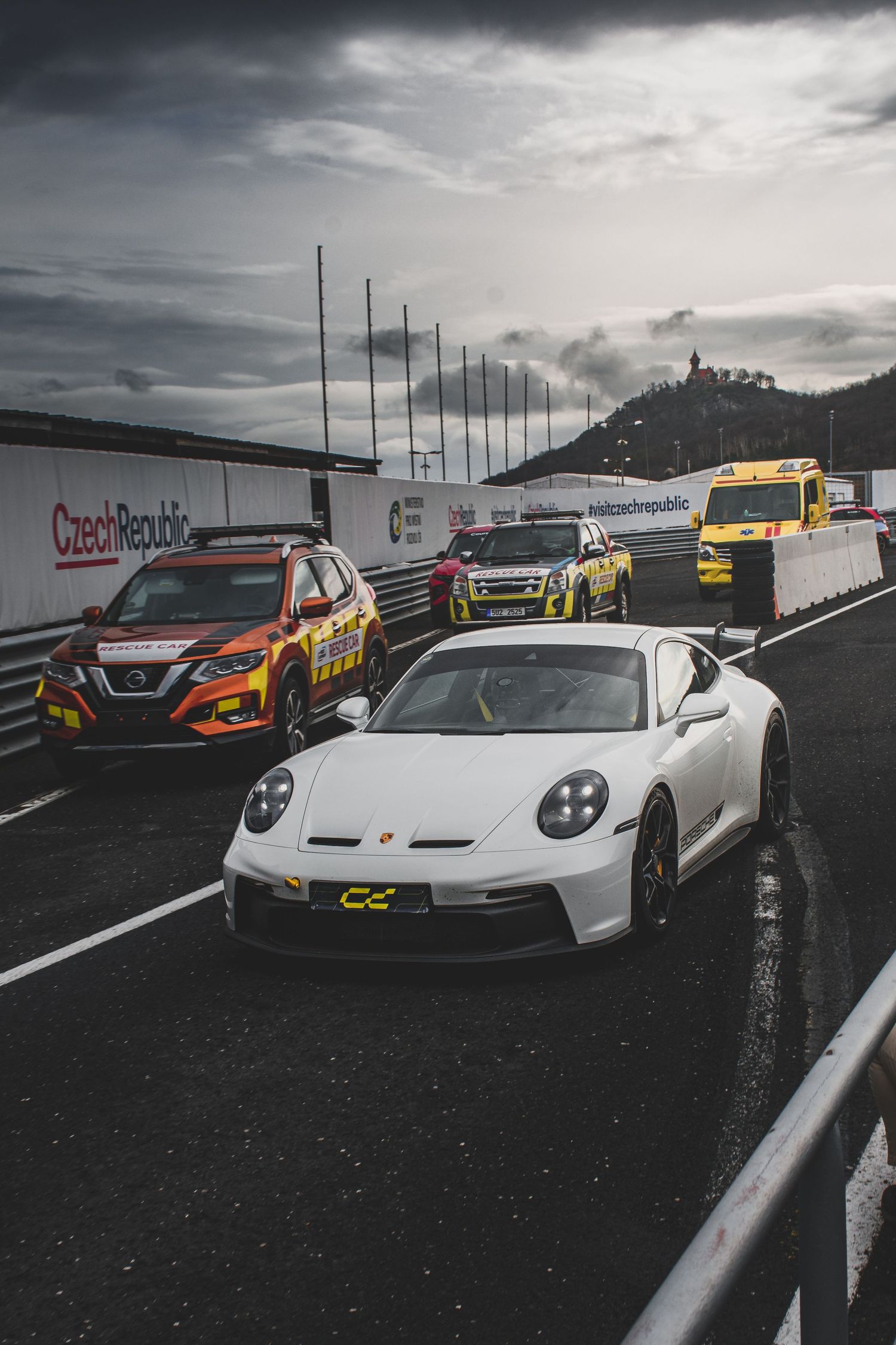 White Porsche 911 GT3 leads safety vehicles on a Czech Republic race circuit.