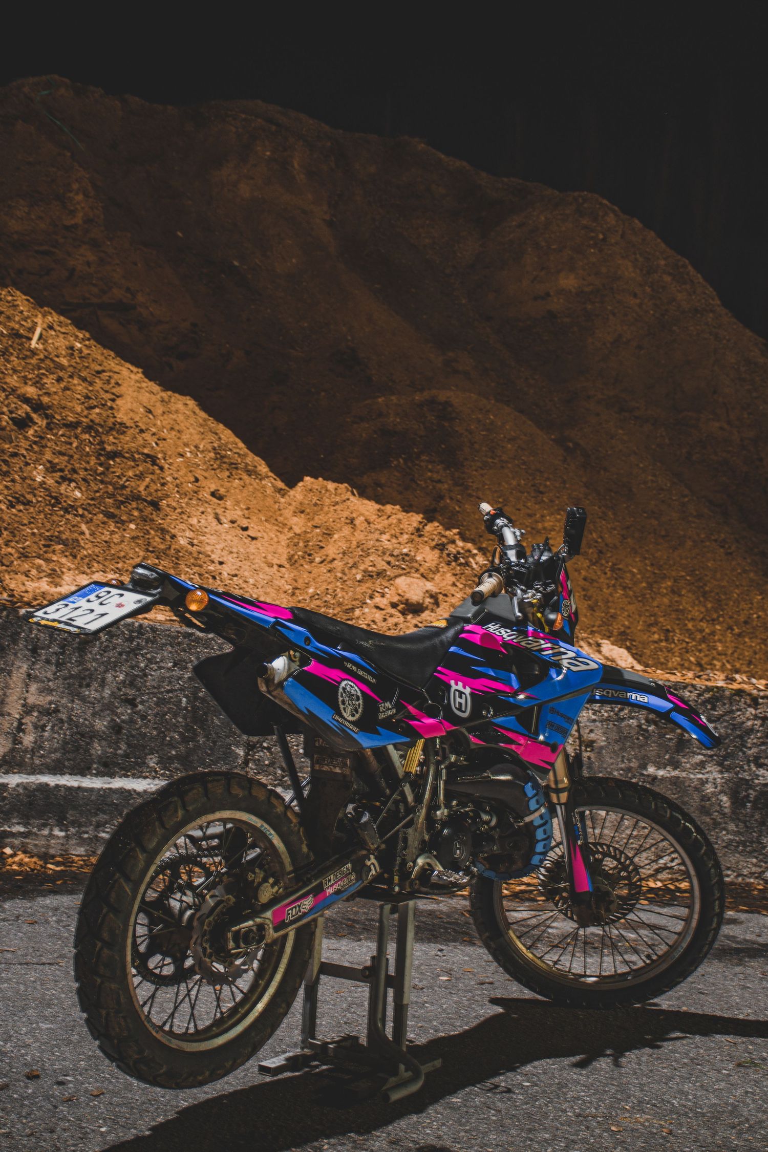 Pink and blue Husqvarna dirt bike parked against a rocky mountain backdrop at night.