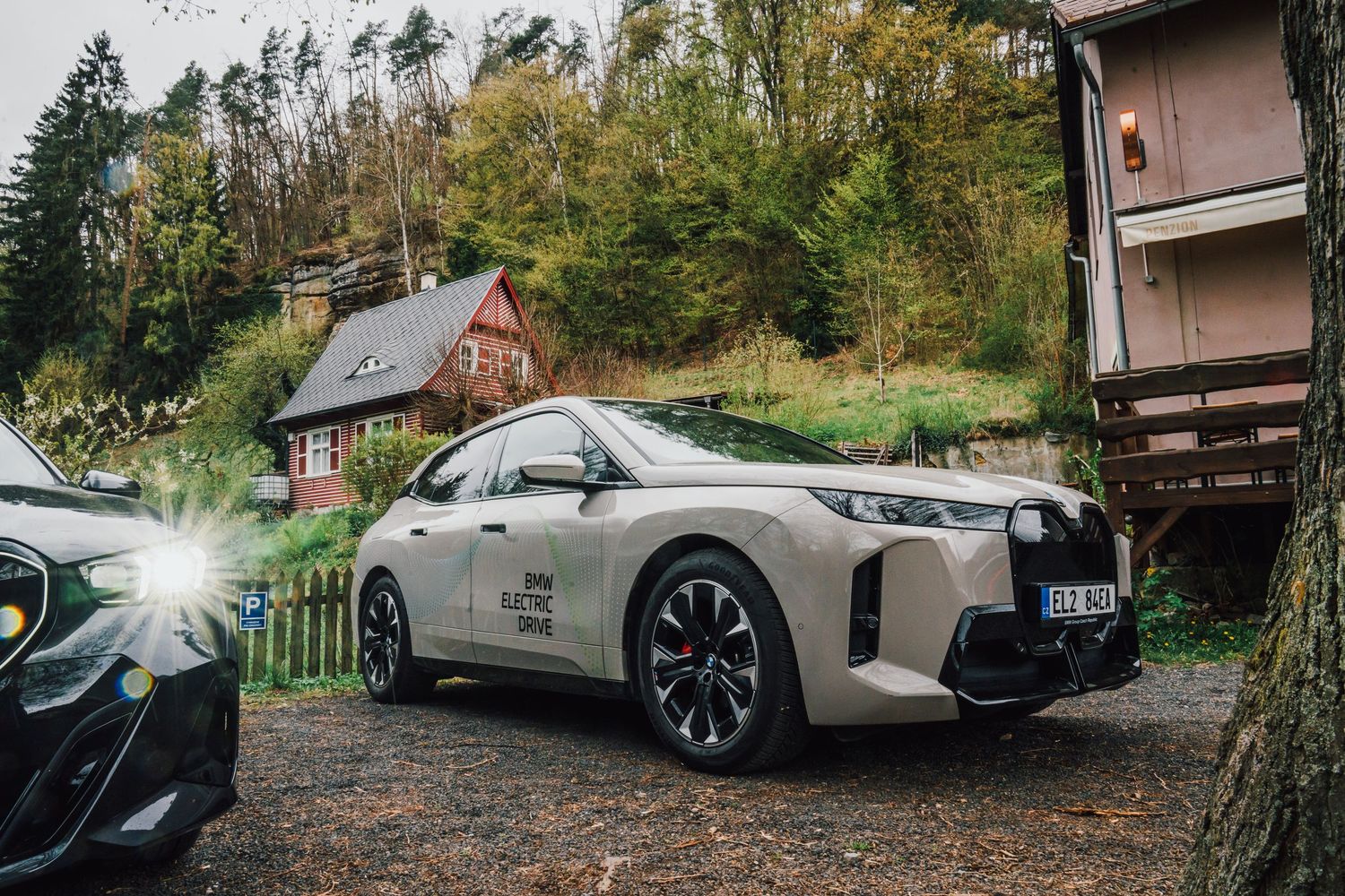 A beige BMW iX electric SUV parked on gravel near a rustic woodland cottage in autumn.