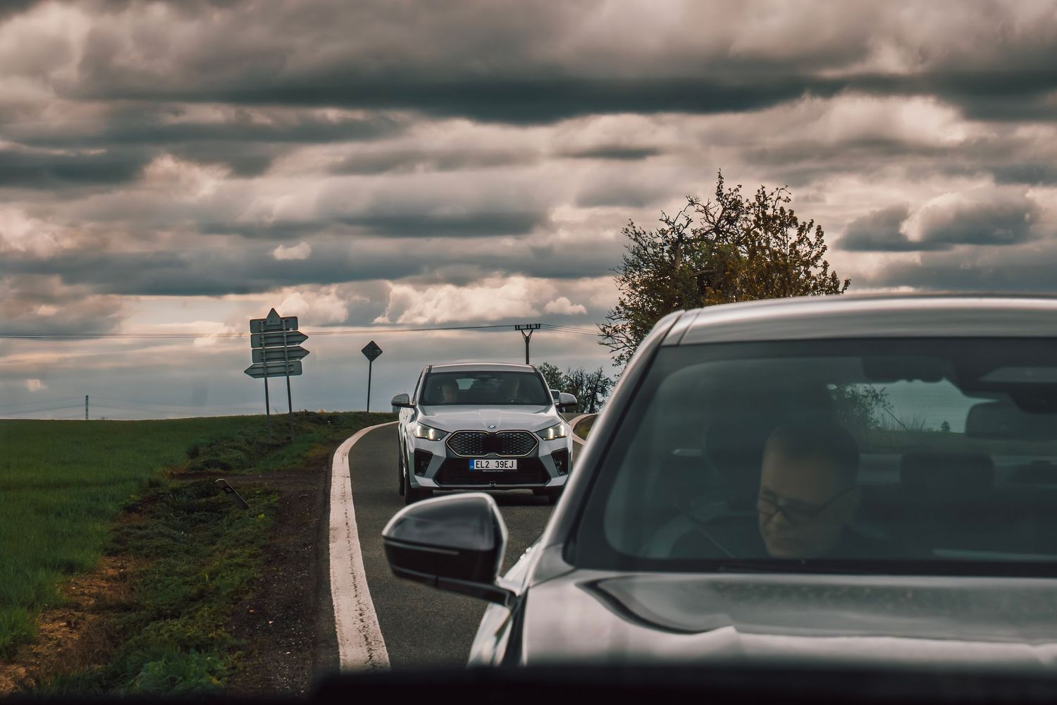 BMW electric cars queued on a rural road with overcast skies, viewed from inside a following vehicle.