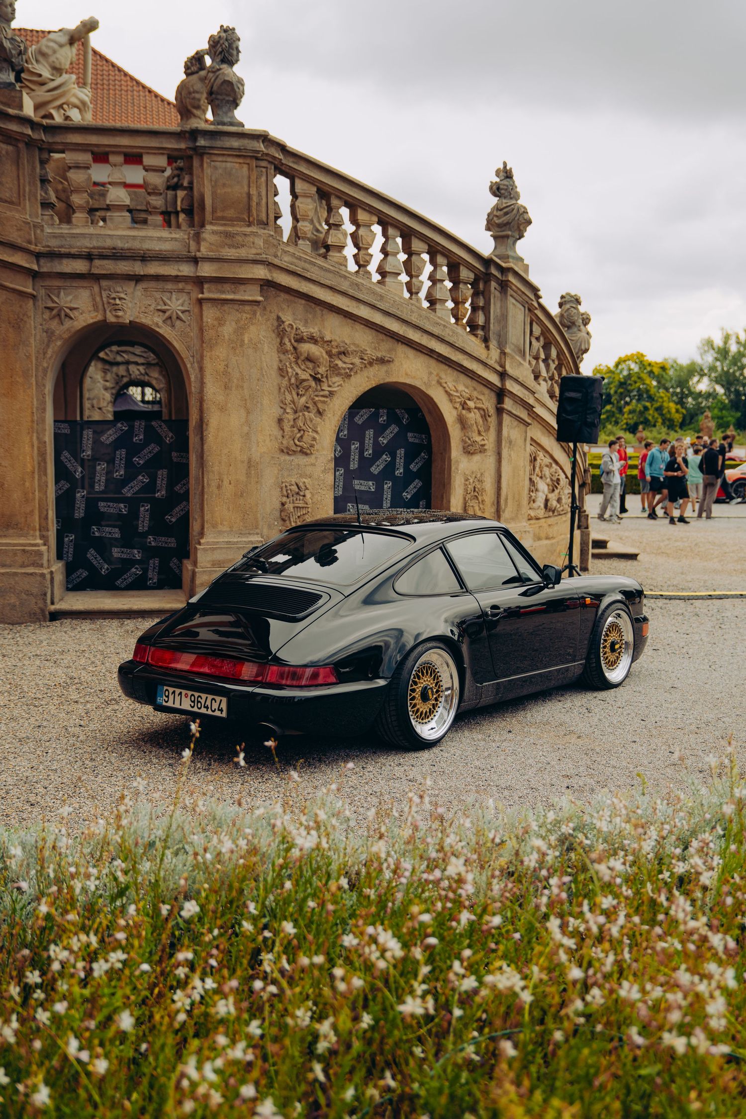 Classic black Porsche 911 with gold BBS wheels parked on gravel in front of ornate baroque architecture.