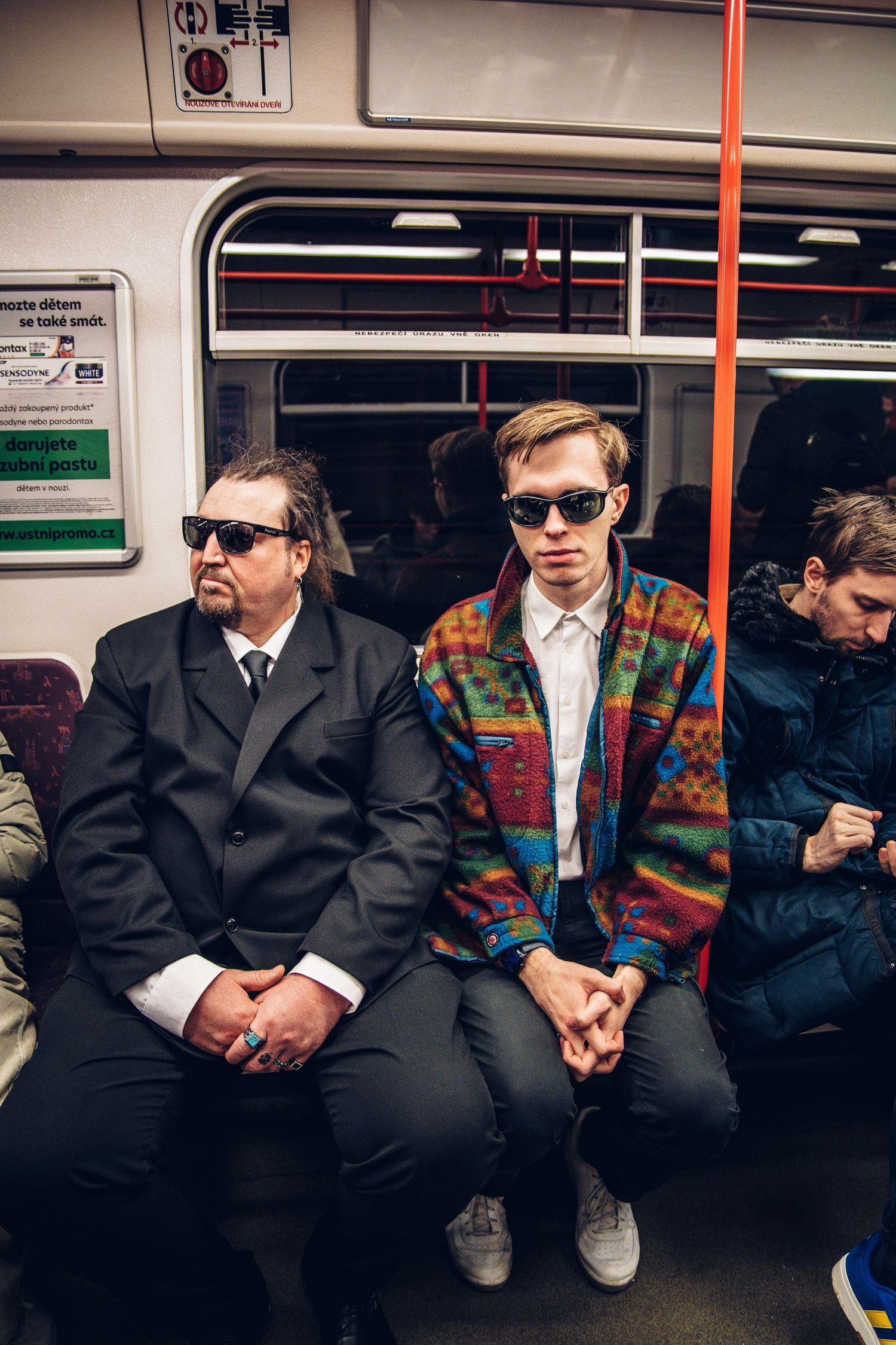 Two men wearing sunglasses sitting on subway train, one in suit and one in colorful jacket.