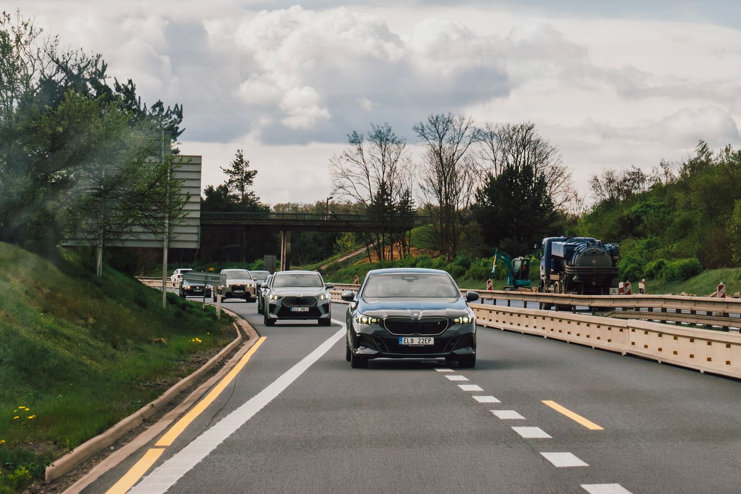 Convoy of BMW vehicles driving on a multi-lane highway under a cloudy overcast sky.