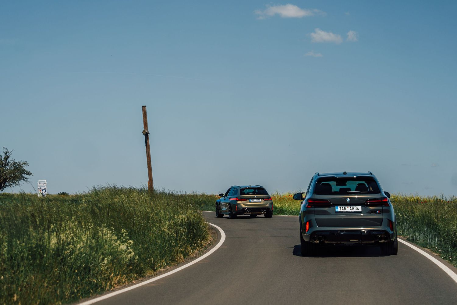 Two BMW M vehicles driving on a rural road surrounded by green fields under a clear blue sky.