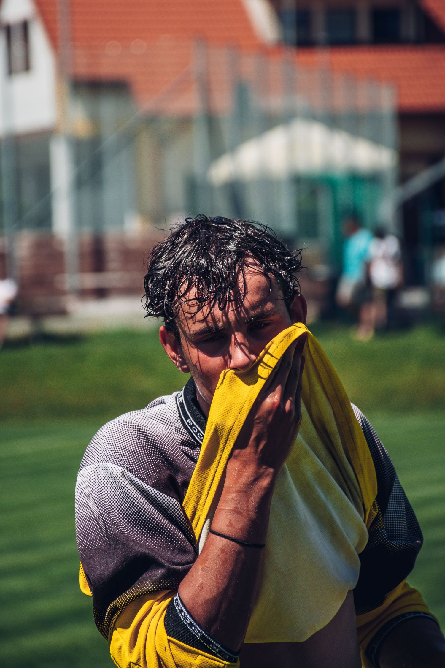 Sweaty male soccer player wiping face with yellow jersey after intense match on outdoor field.