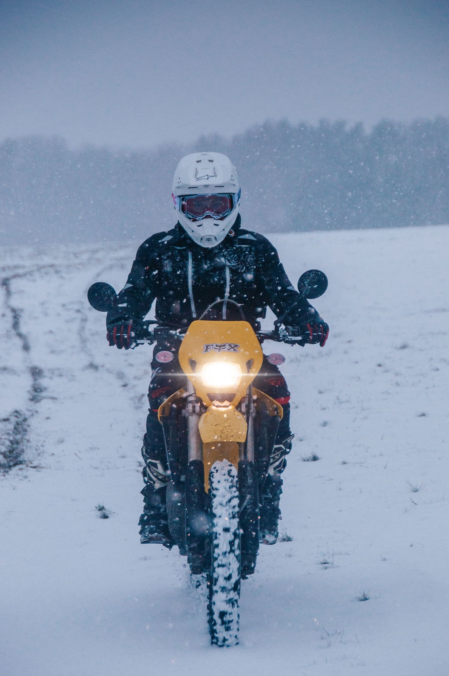 Motocross rider on yellow dirt bike rides through heavy snowstorm with headlight illuminated.
