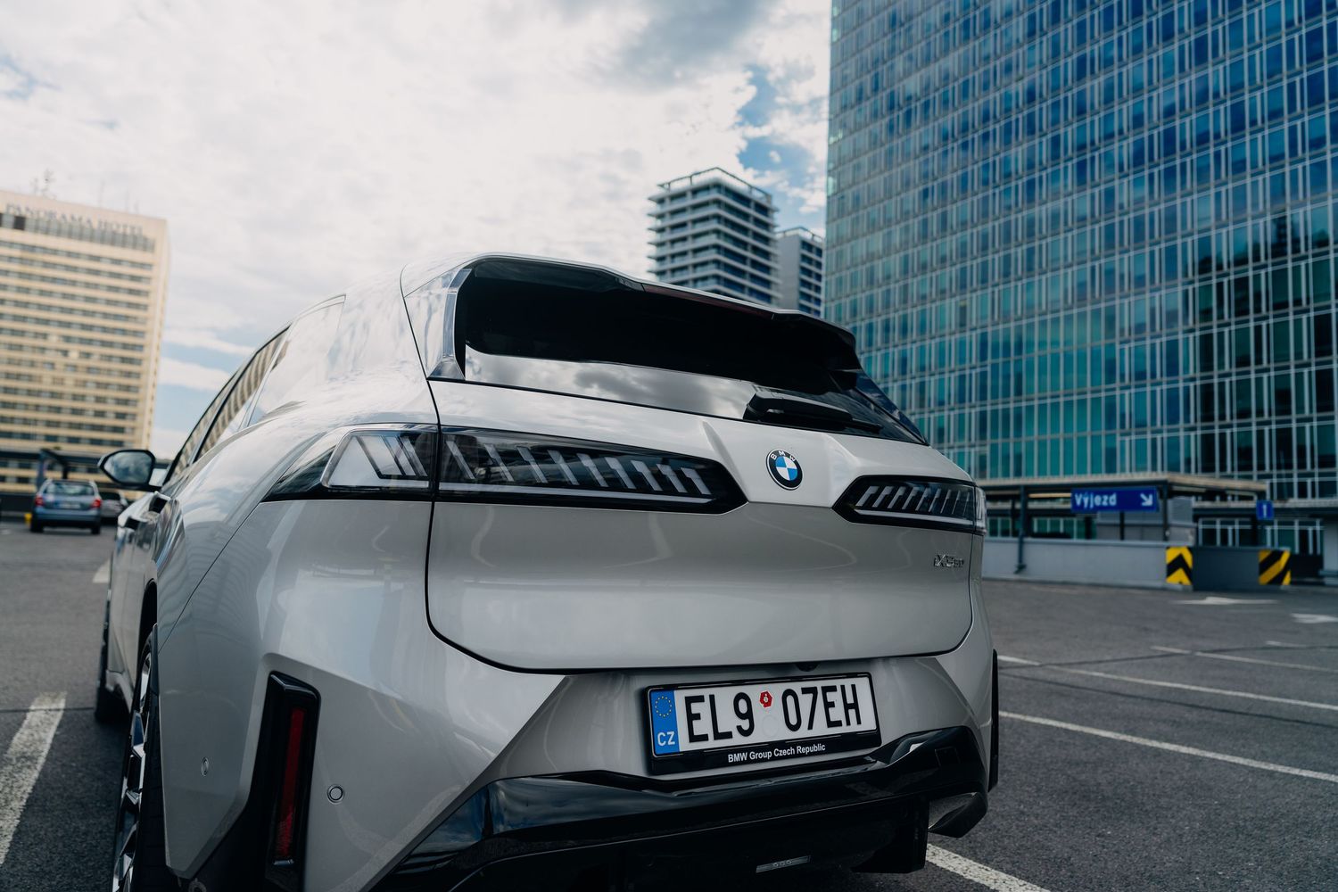 Rear view of a silver BMW iX electric SUV parked in an urban setting with modern glass buildings in the background.