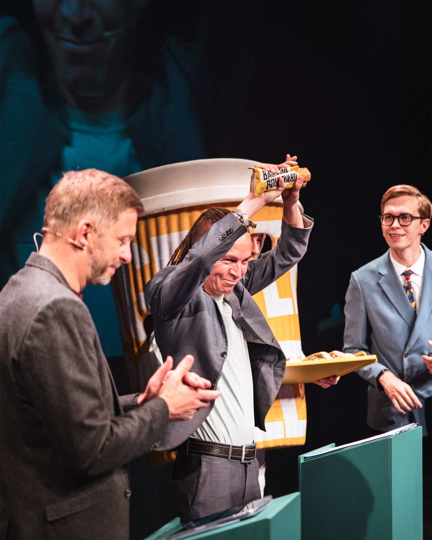 Man raising a trophy at an award ceremony flanked by two men on a teal-decorated stage.