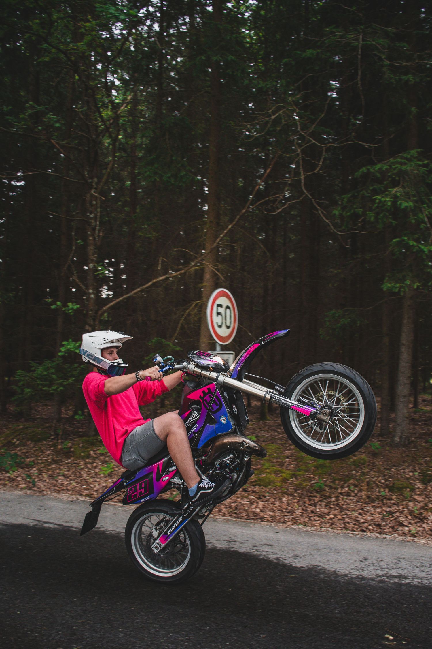 Daring rider performs wheelie on purple dirt bike near 50mph speed sign in forest.