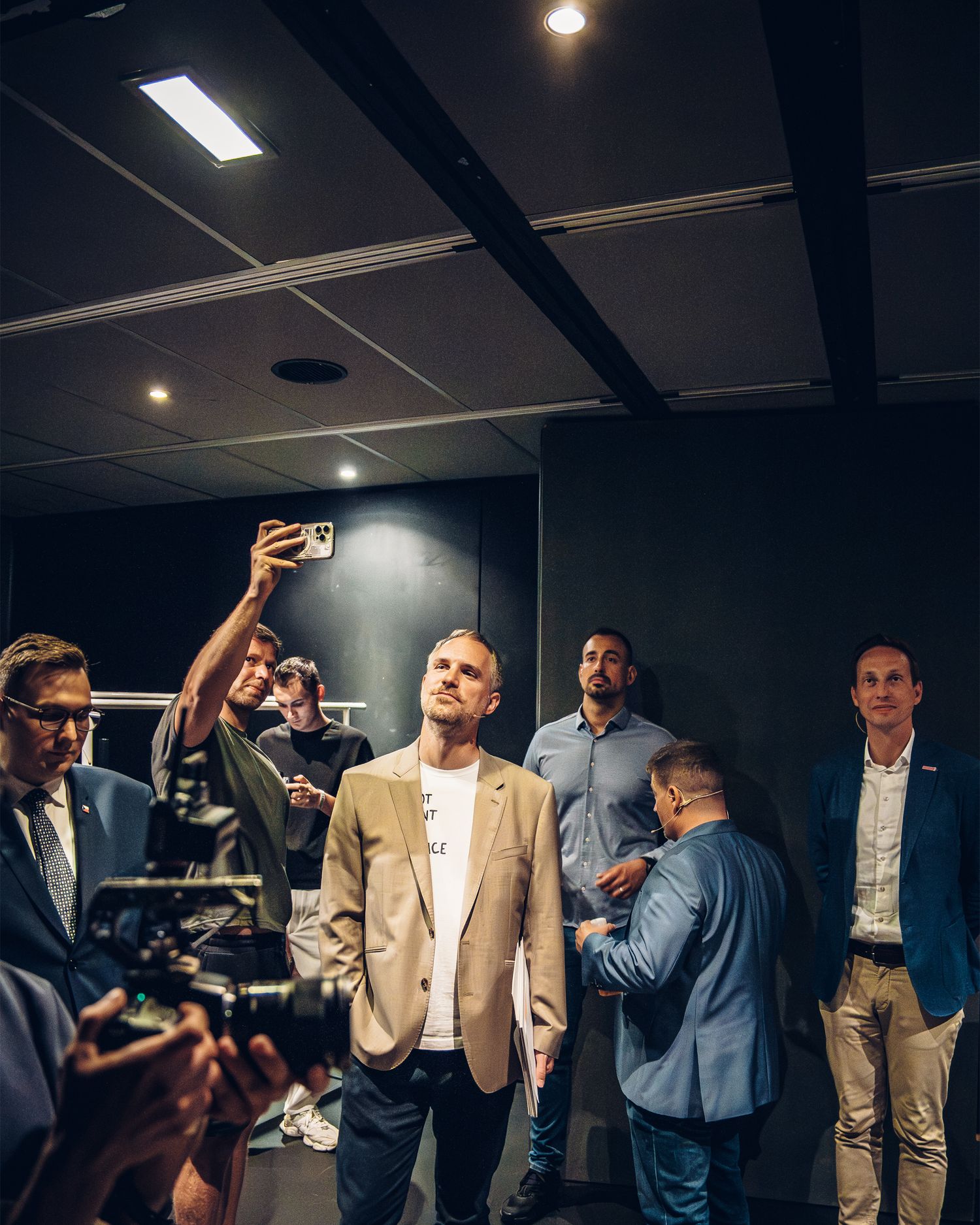 Group of men in a dark room, one taking a selfie while others stand nearby at a professional event.
