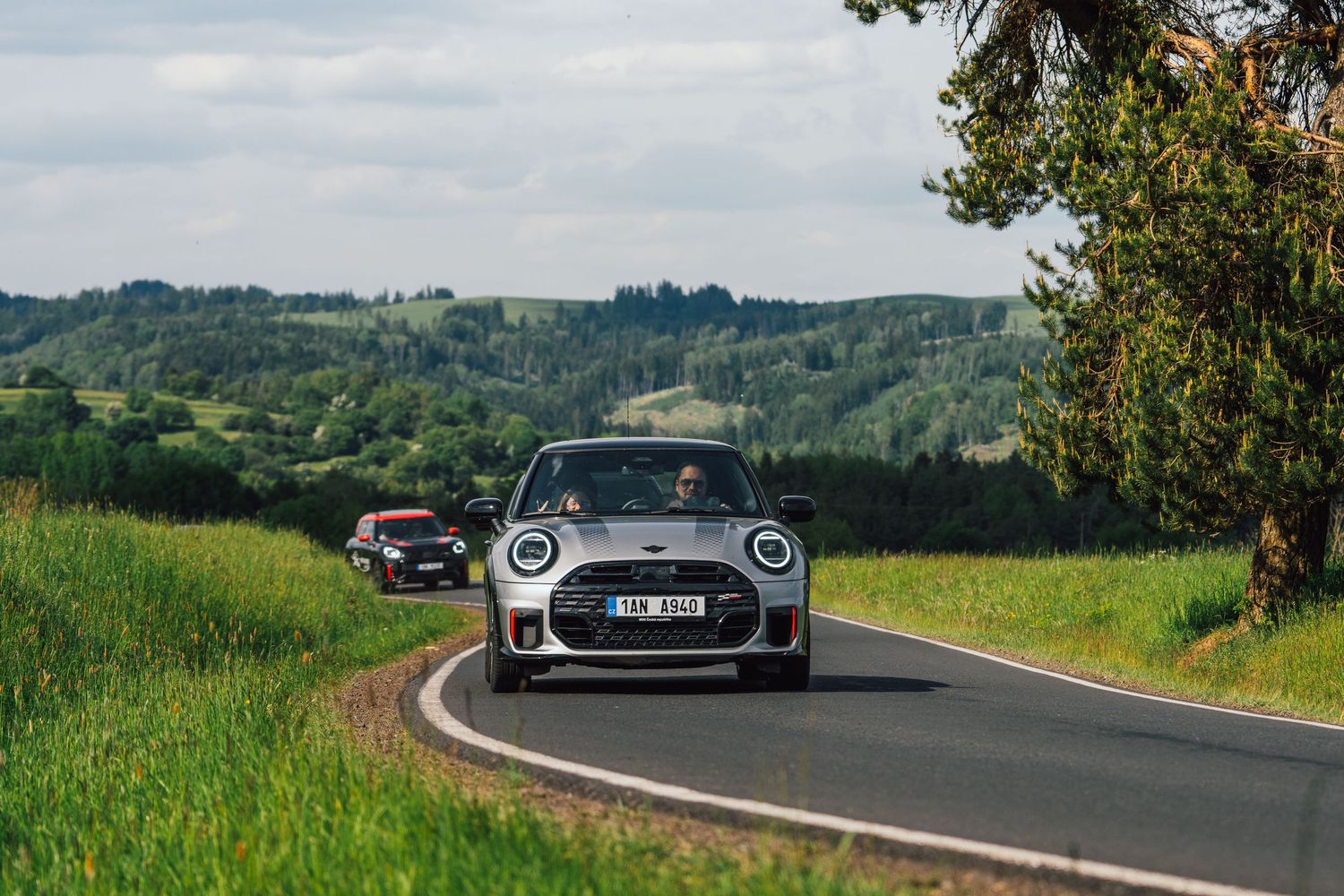 Two MINI Cooper hatchbacks driving on a scenic countryside road with rolling green hills.