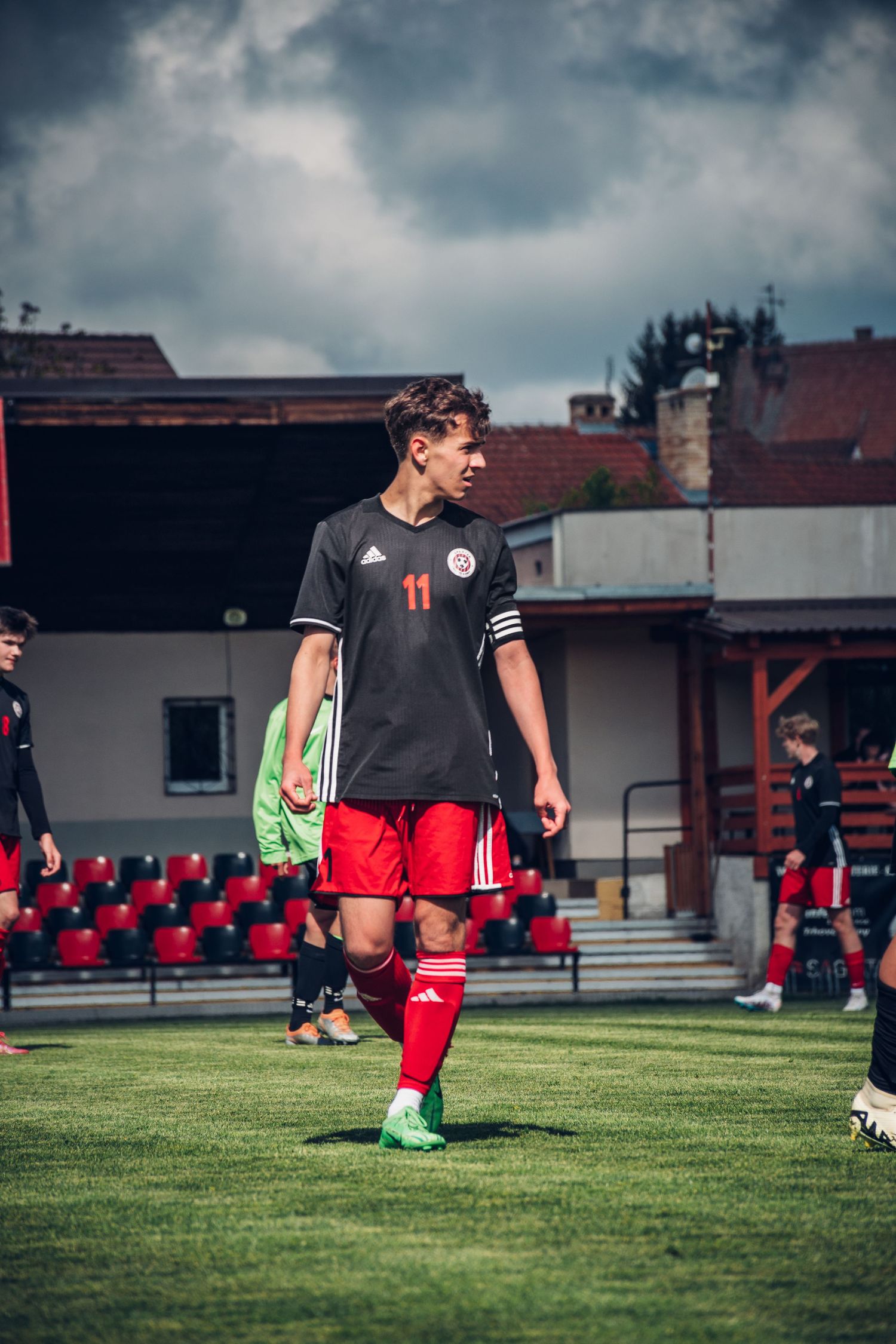 Young soccer player wearing number 11 black Adidas jersey and red shorts walks on a green field during a game.