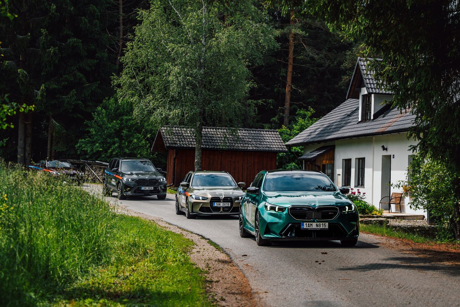 Convoy of BMW SUVs and a green BMW X5 M driving through a quiet village road lined with trees.