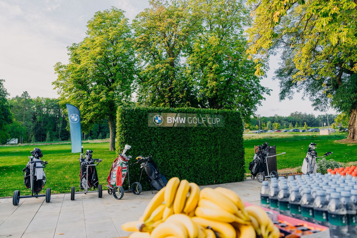 Golf bags lined up at BMW Golf Cup event with bananas and water bottles in foreground, green hedge backdrop.