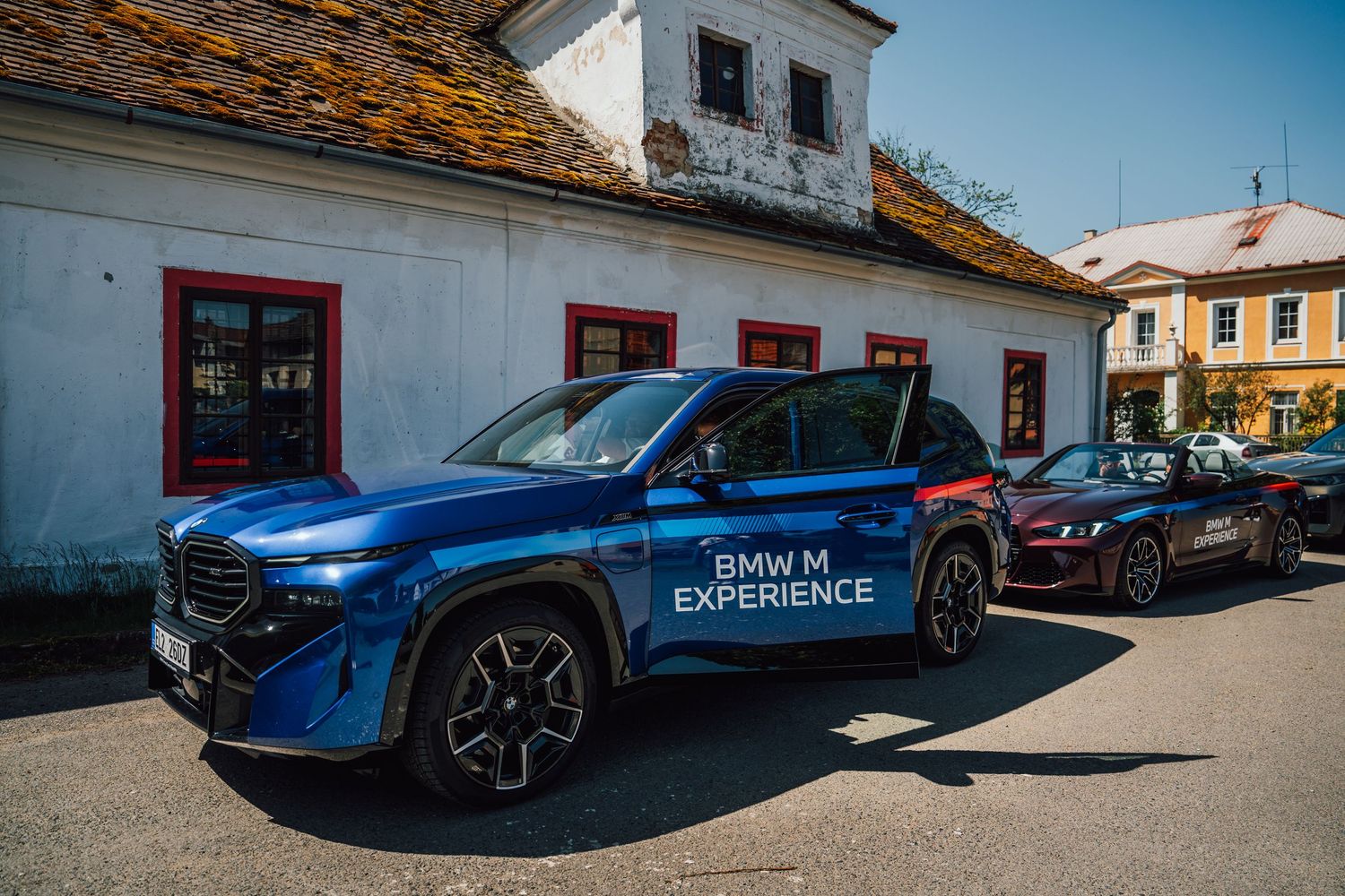 Blue BMW XM M Experience SUV parked in front of a historic white building in a village.