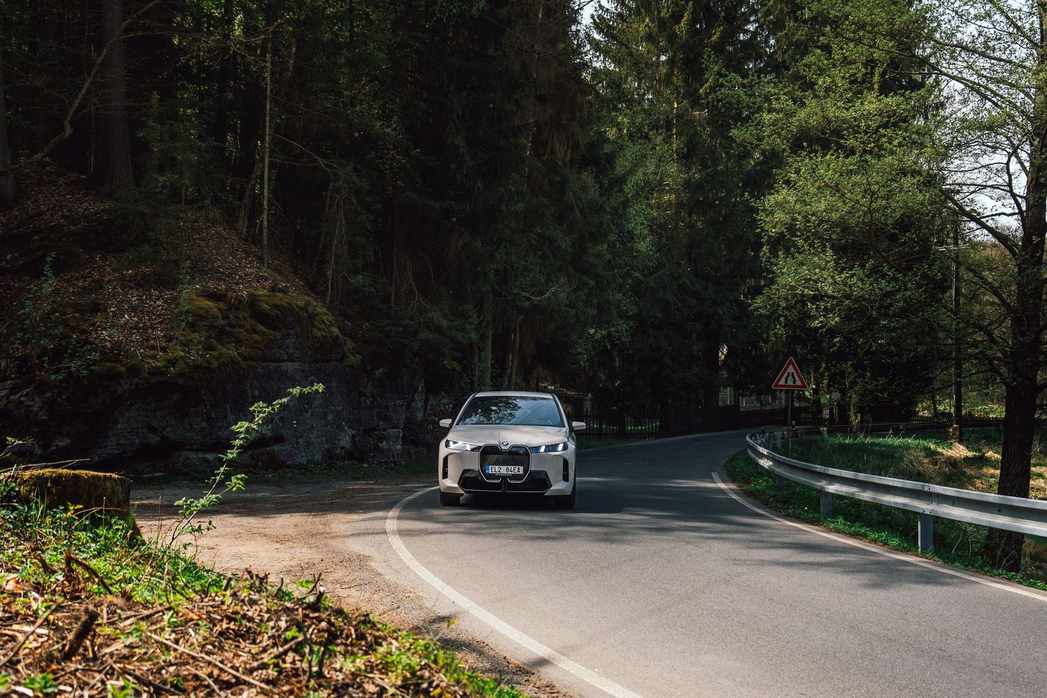 BMW SUV driving along a narrow forest road surrounded by tall pine trees in natural daylight.