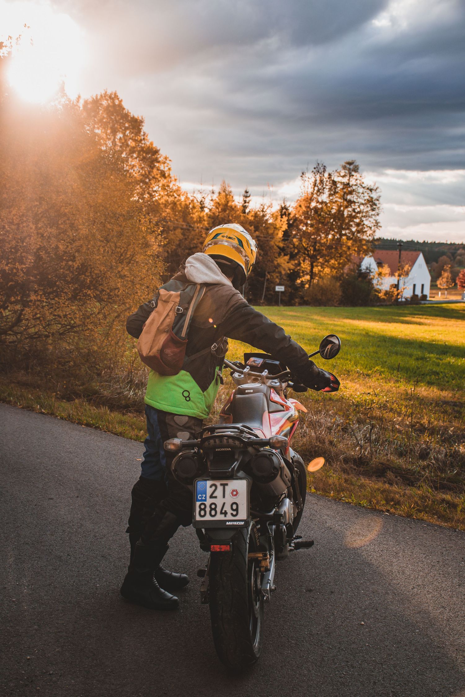 Motorcyclist with yellow helmet and backpack on road at golden sunset with countryside background.