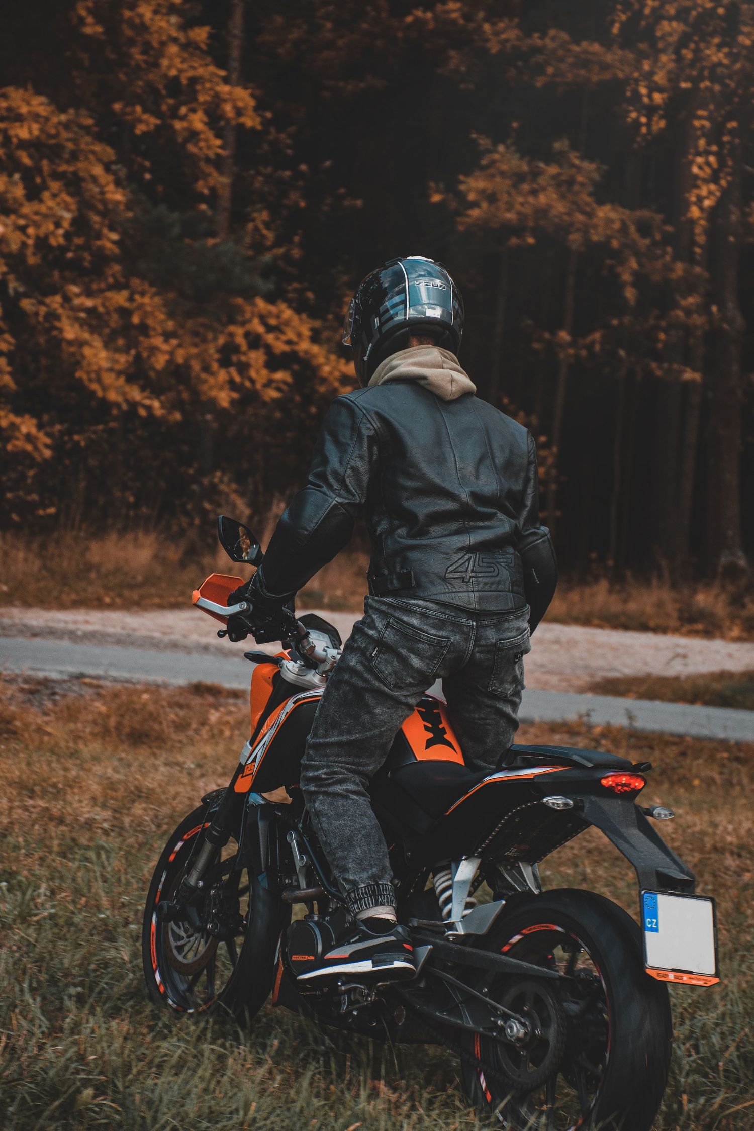 Motorcyclist in black leather gear and helmet sitting on an orange KTM motorcycle in an autumn forest.