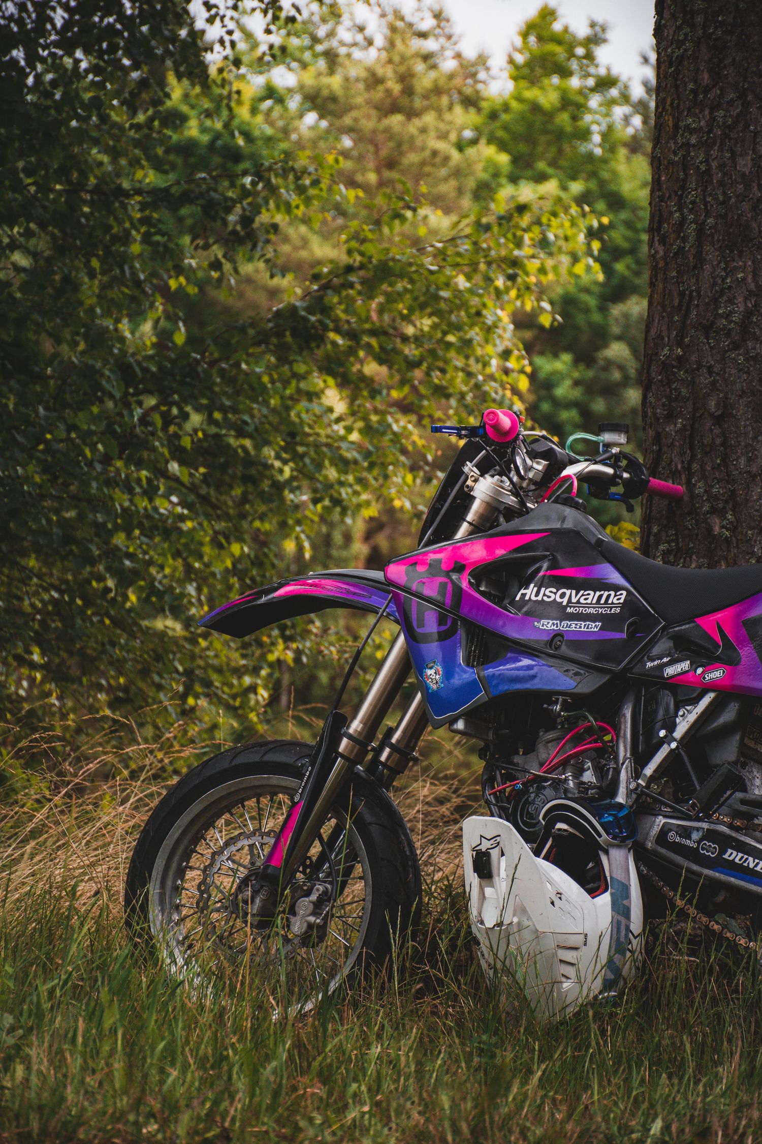 Purple and pink Husqvarna dirt bike leaning against a tree in a lush green forest setting.