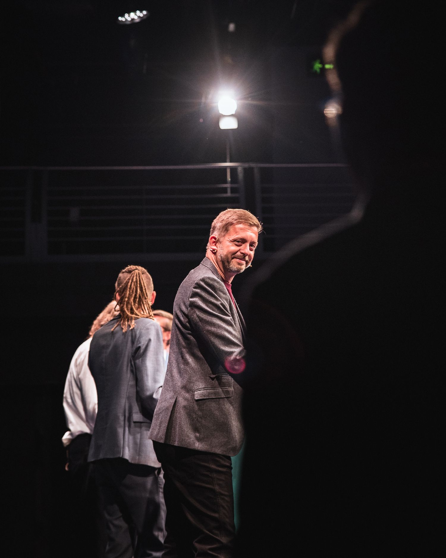Man in grey blazer smiling on a dark stage with spotlight and another person standing behind him.