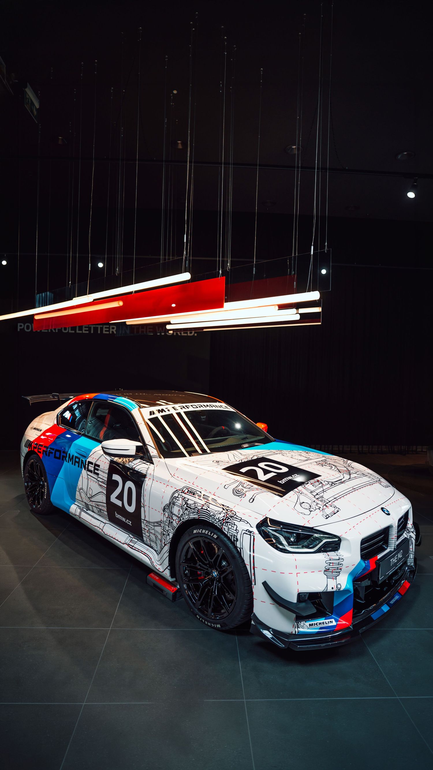 BMW M2 GT4 race car number 20 with colorful livery displayed indoors under dramatic lighting.