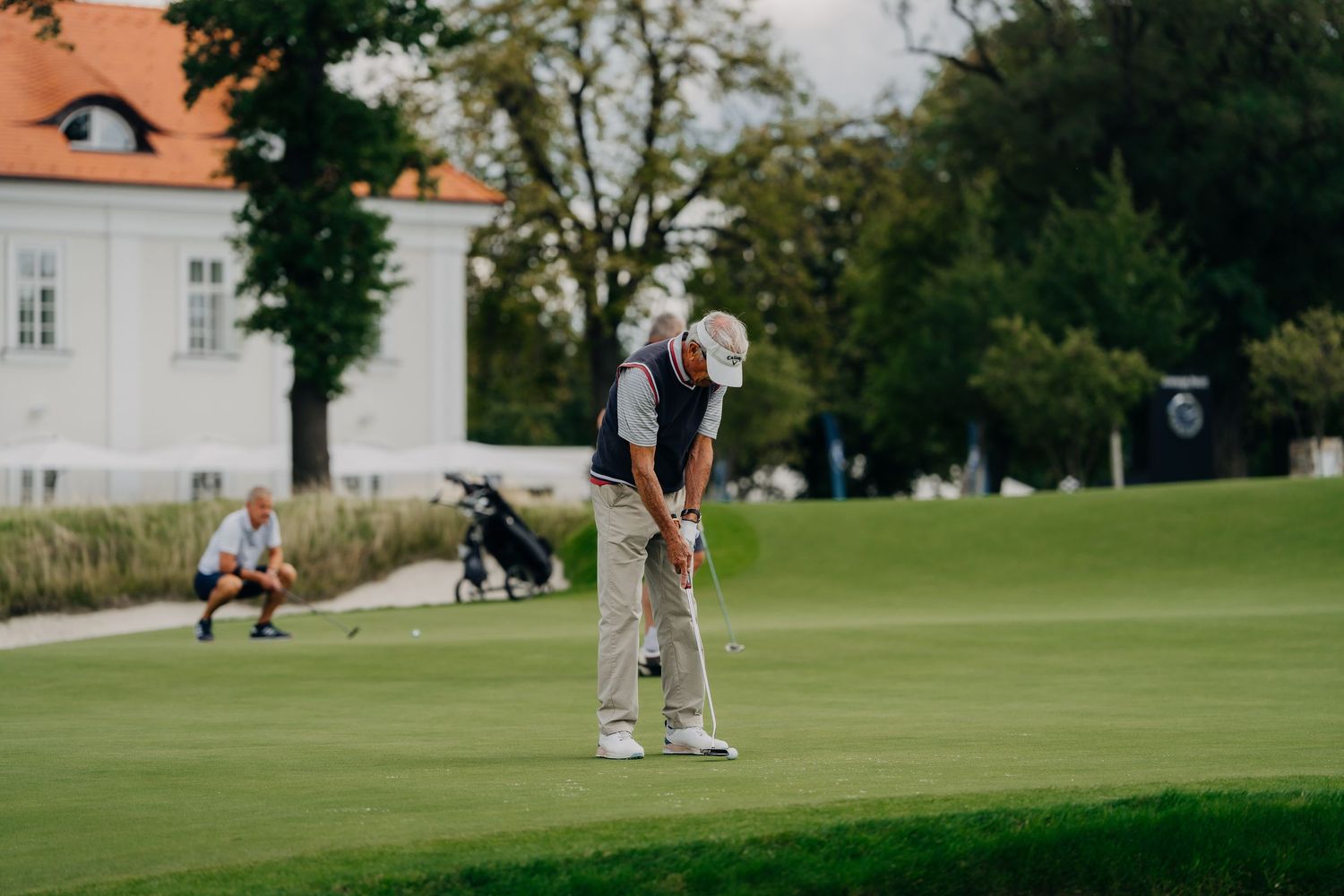 Senior golfer putting on green at BMW Golf Cup with historic white manor house in background.