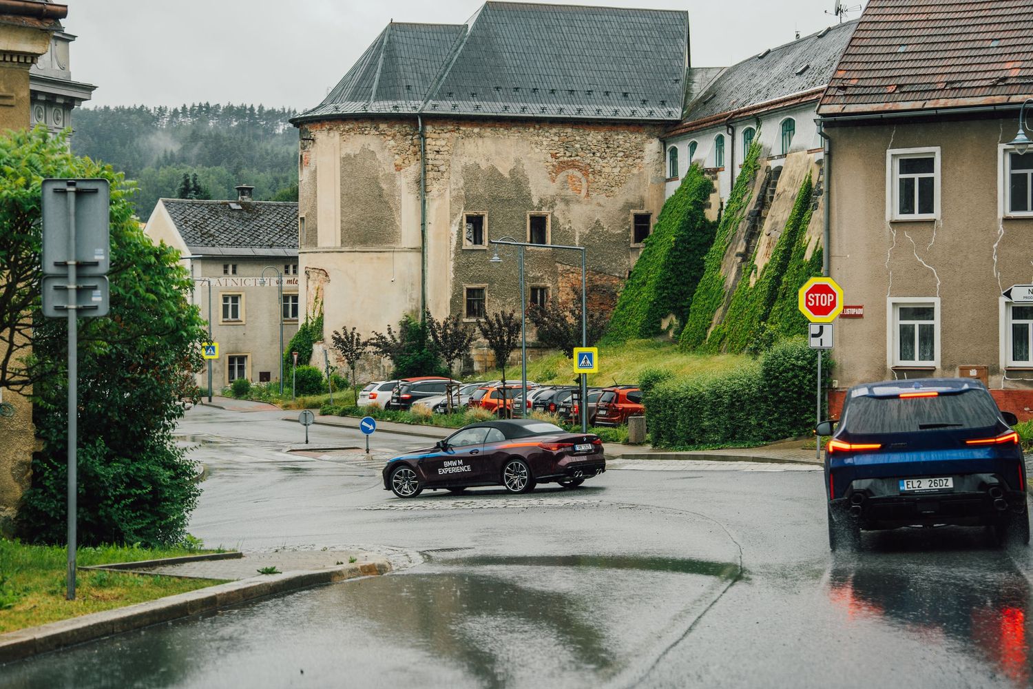 BMW M Experience cars driving through a rainy Central European village with historic church and traditional architecture.