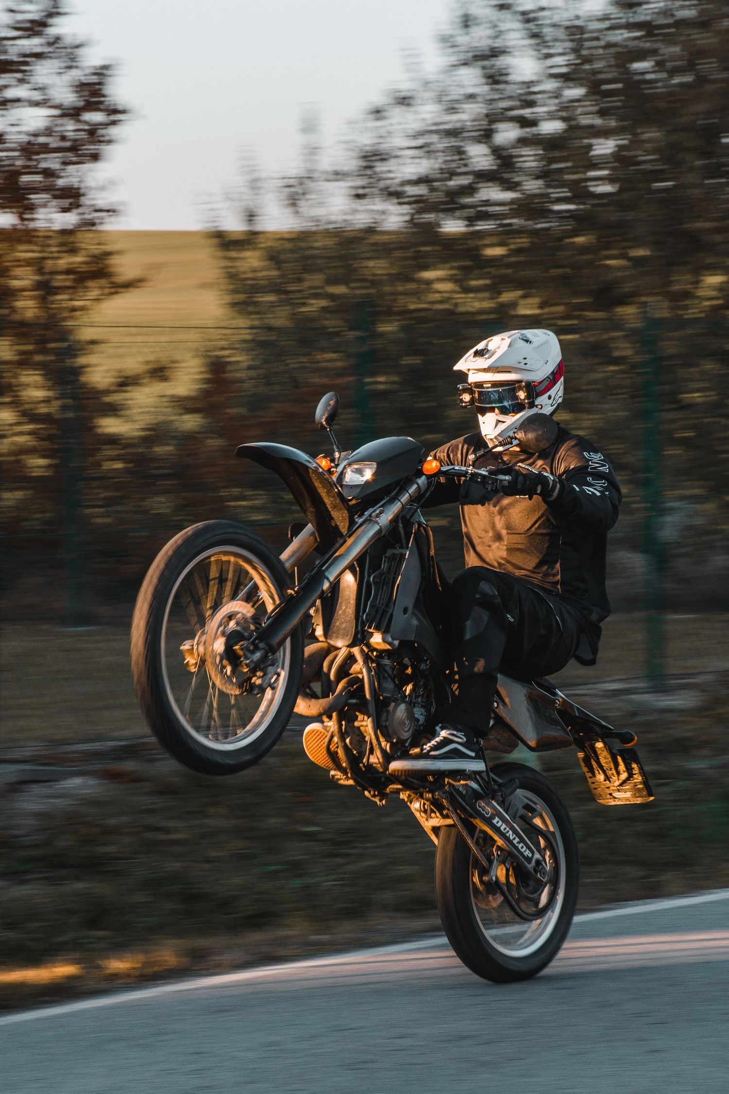 Motorcyclist doing wheelie on orange and black supermoto bike on rural road at sunset.