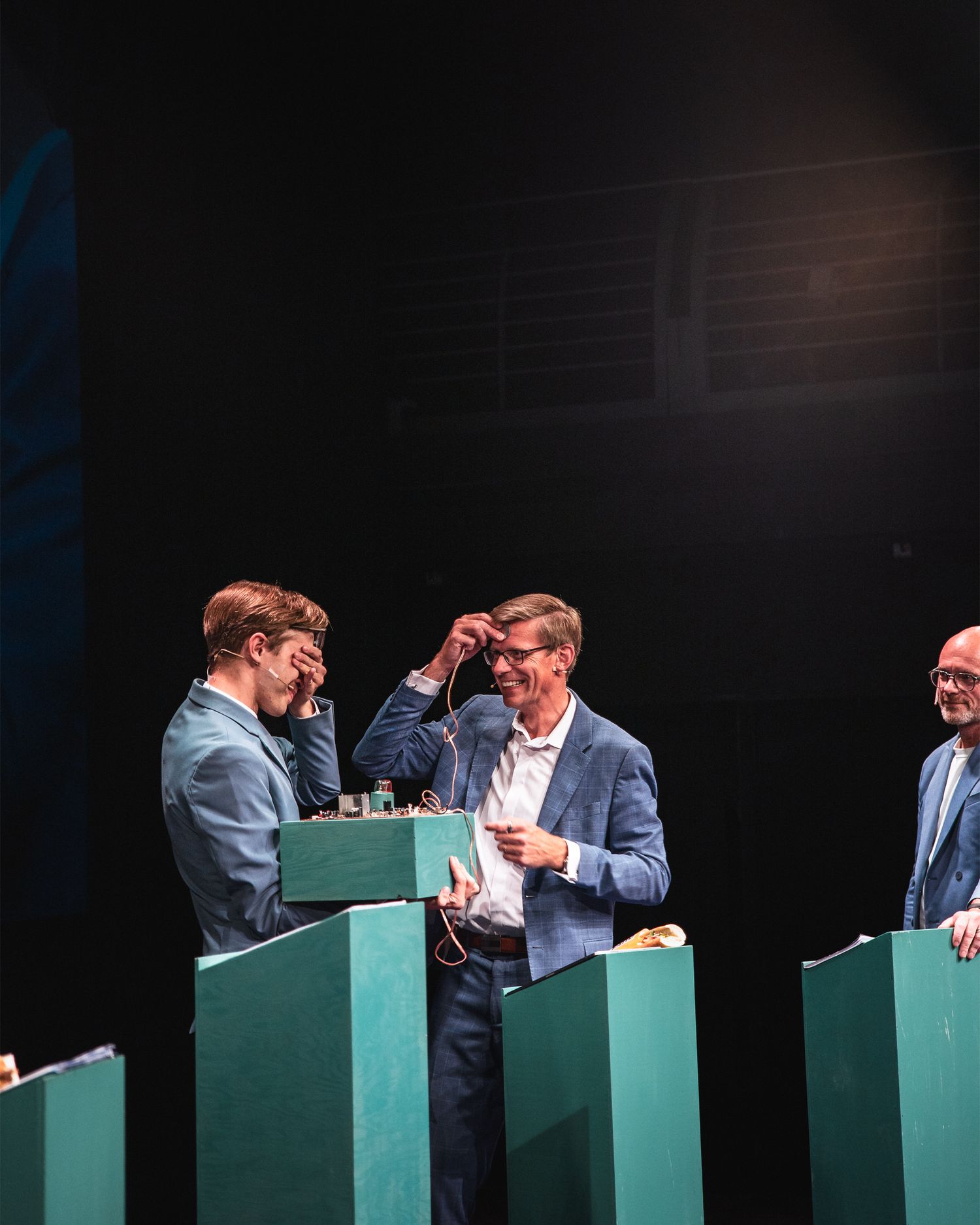 Three men in suits interacting behind teal podiums on dark theater stage during live event.
