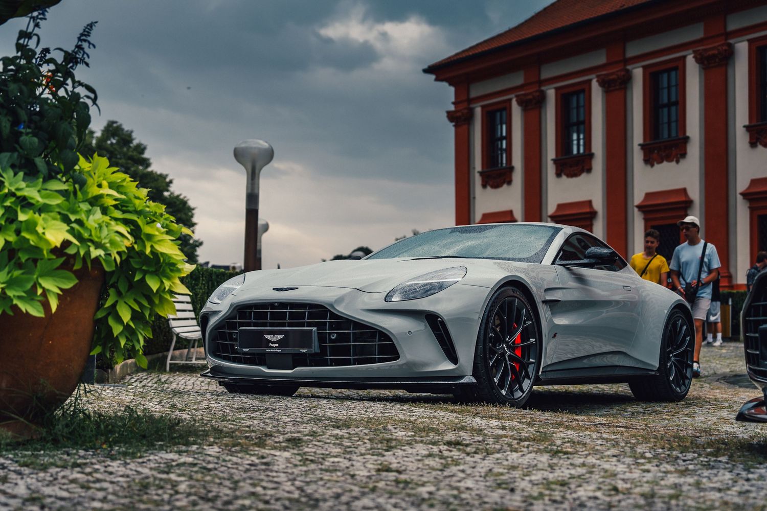 Silver Aston Martin sports car parked on cobblestones before a red-roofed historic building under dramatic cloudy skies.