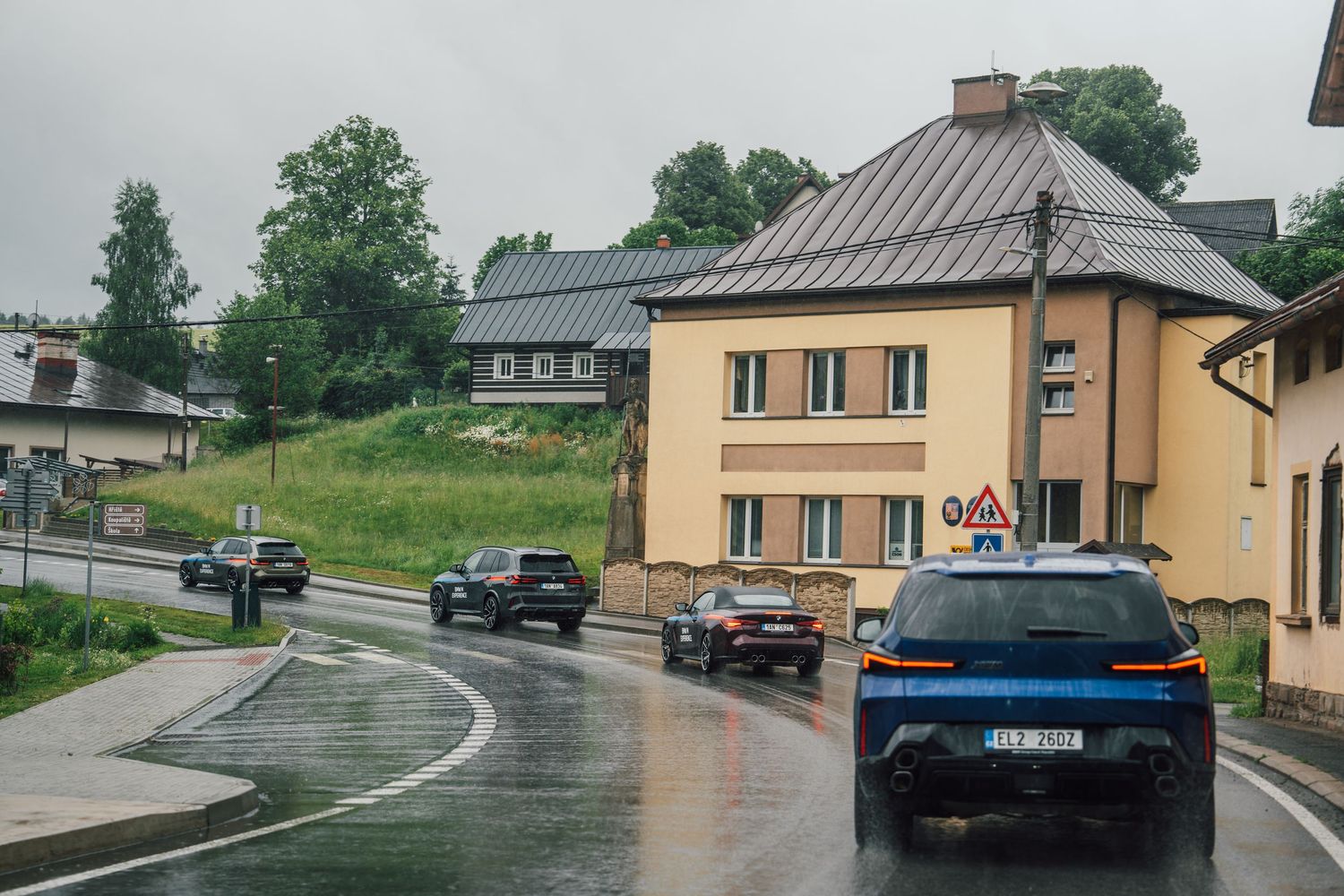 A convoy of BMW cars driving through a wet village street on a rainy day in Central Europe.