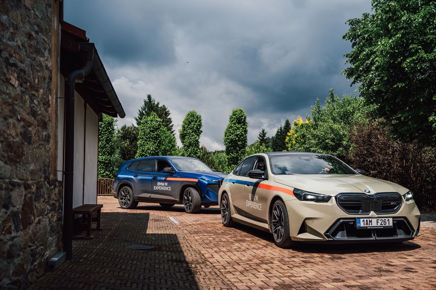 BMW M Experience gold sedan and blue XM SUV parked on brick driveway at rural location.