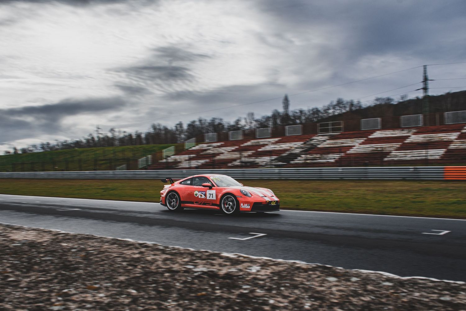 Orange Porsche 911 race car speeding on a wet racing circuit under cloudy skies.