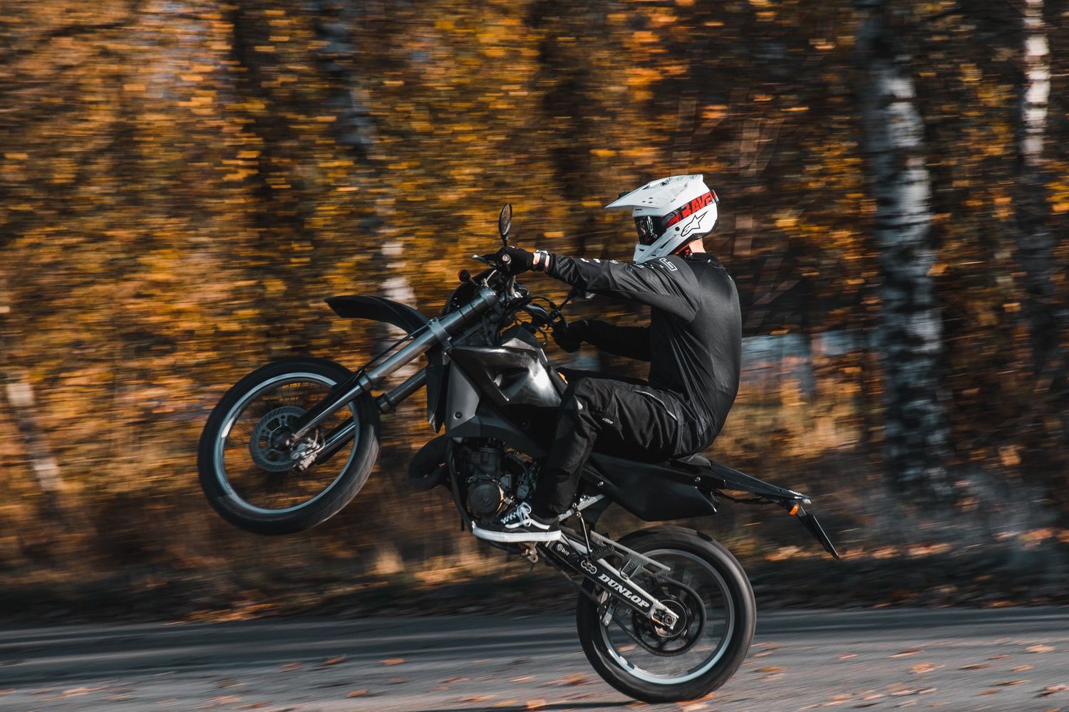 Motorcyclist performing wheelie on black supermoto bike on autumn tree-lined road with motion blur.