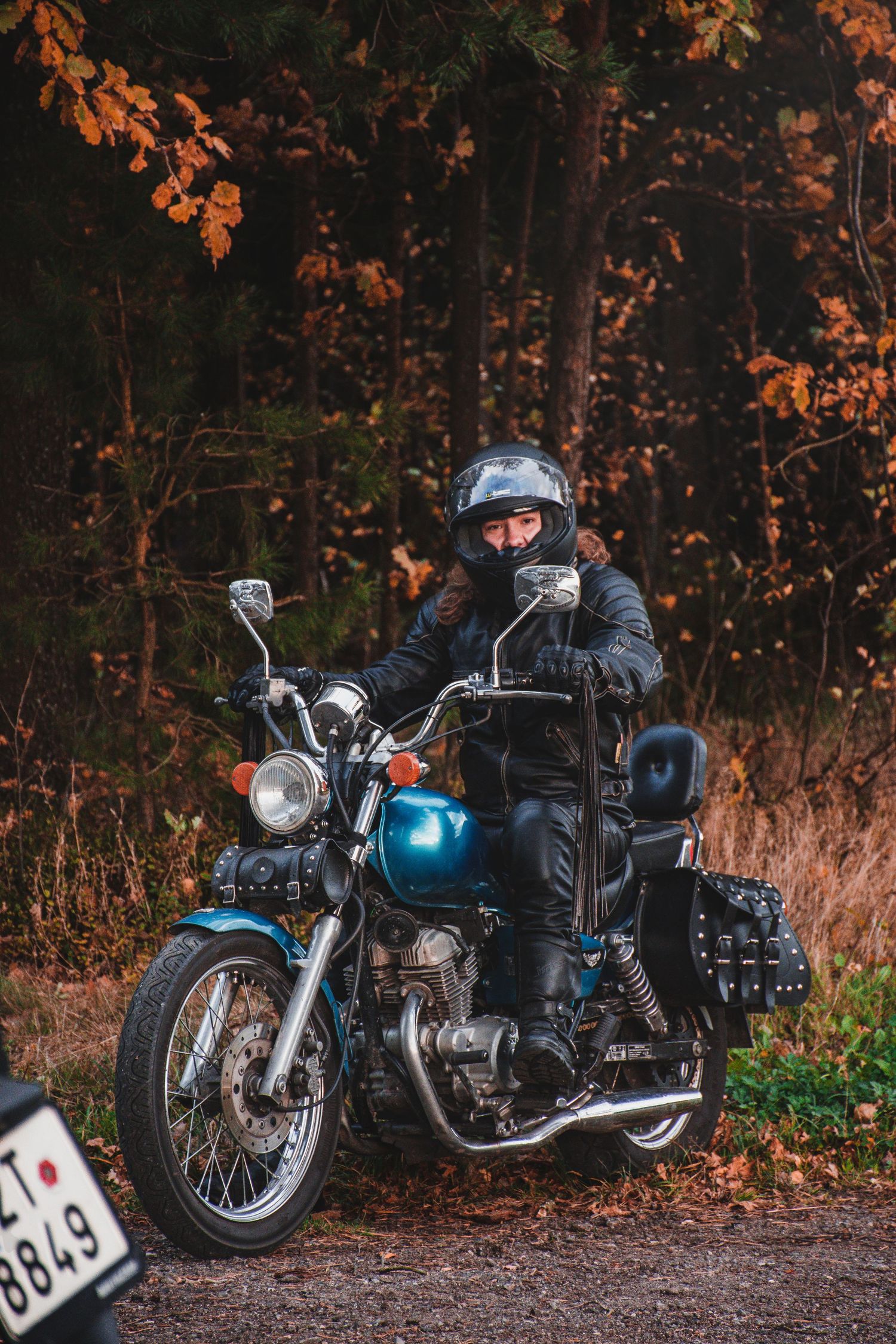 Female motorcyclist in black leather jacket and helmet posing on a vintage blue motorcycle in autumn forest.