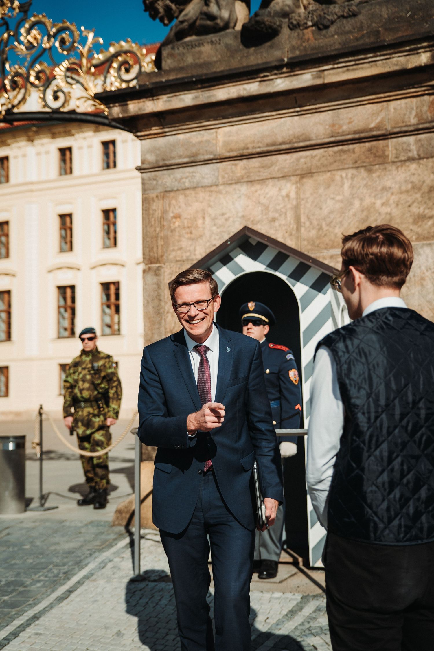 Two men in suits greet each other outside Prague Castle with a guard nearby.