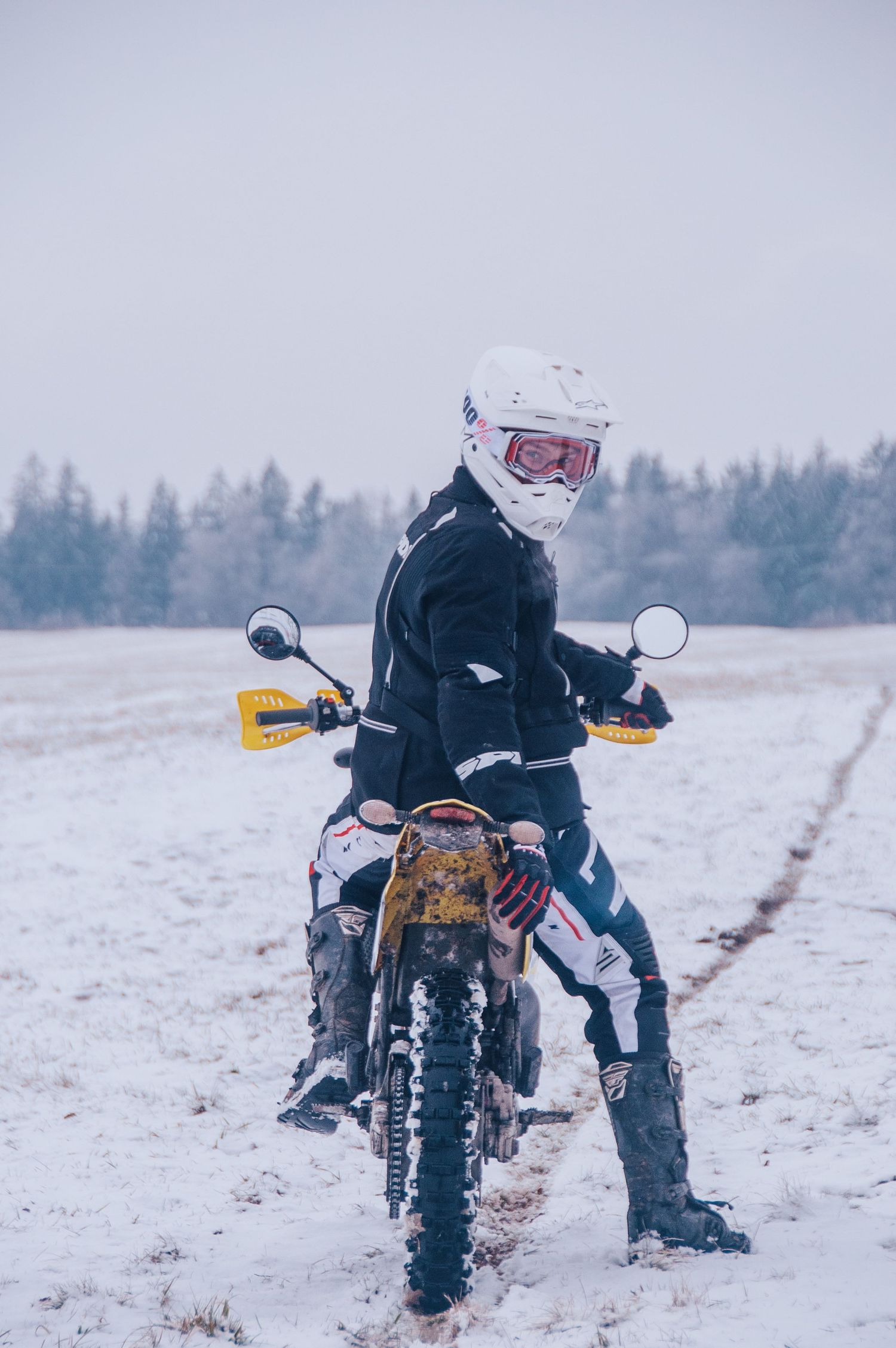 Female motocross rider poses on muddy dirt bike in snowy open field wearing white helmet.