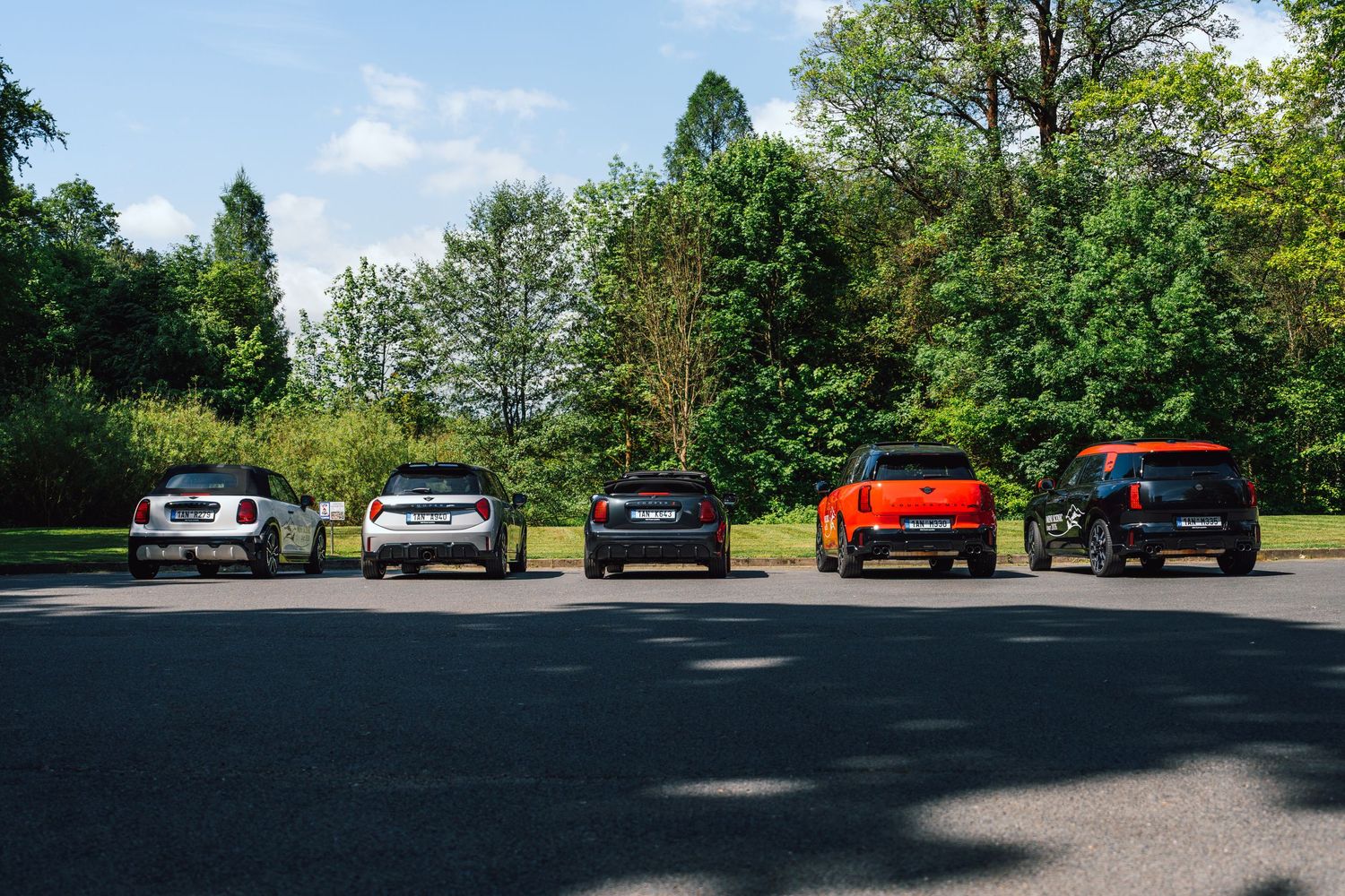 Five MINI Cooper models parked rear-facing in a sunny parking lot surrounded by green trees.