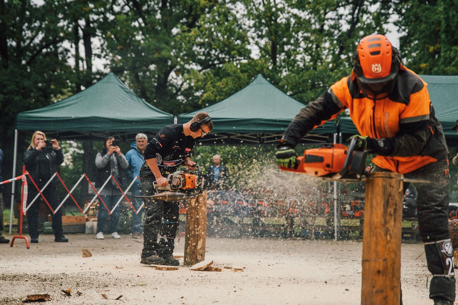 Two competitors use chainsaws simultaneously on wooden posts at outdoor lumberjack sports competition.