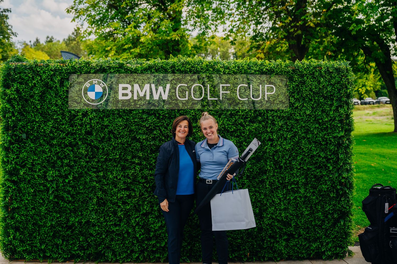 Two smiling participants posing in front of BMW Golf Cup hedge sign at golf tournament event.